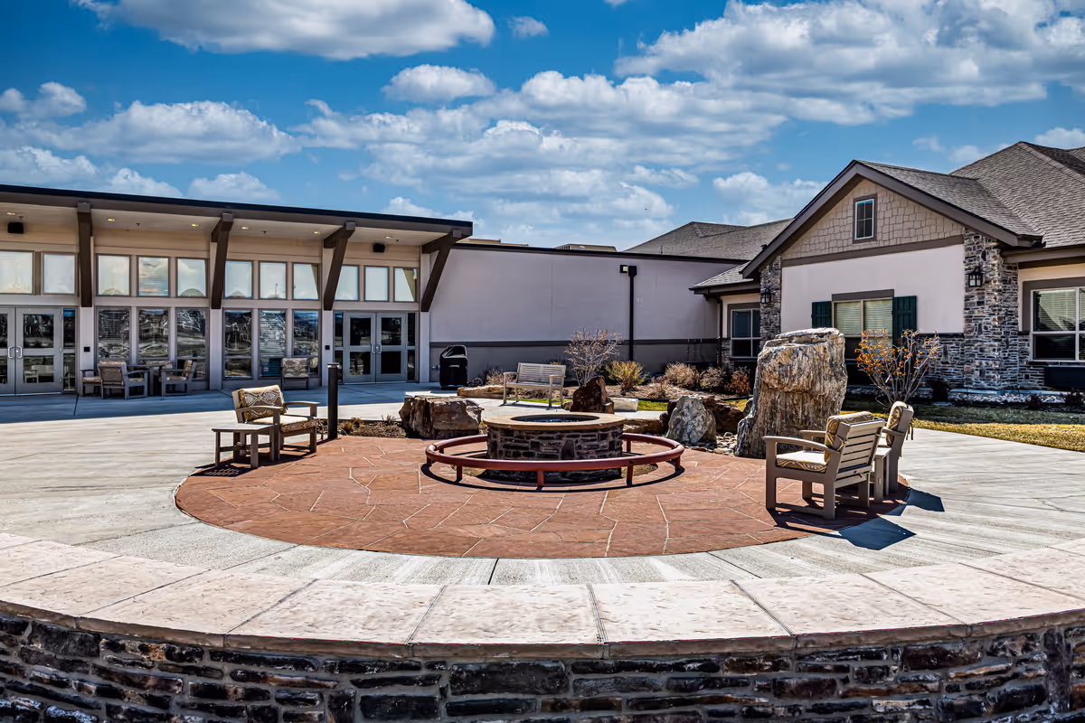 Outdoor patio area at The Peaks at Old Laramie Trail featuring a circular stone fire pit surrounded by chairs, with a building in the background under a partly cloudy sky.