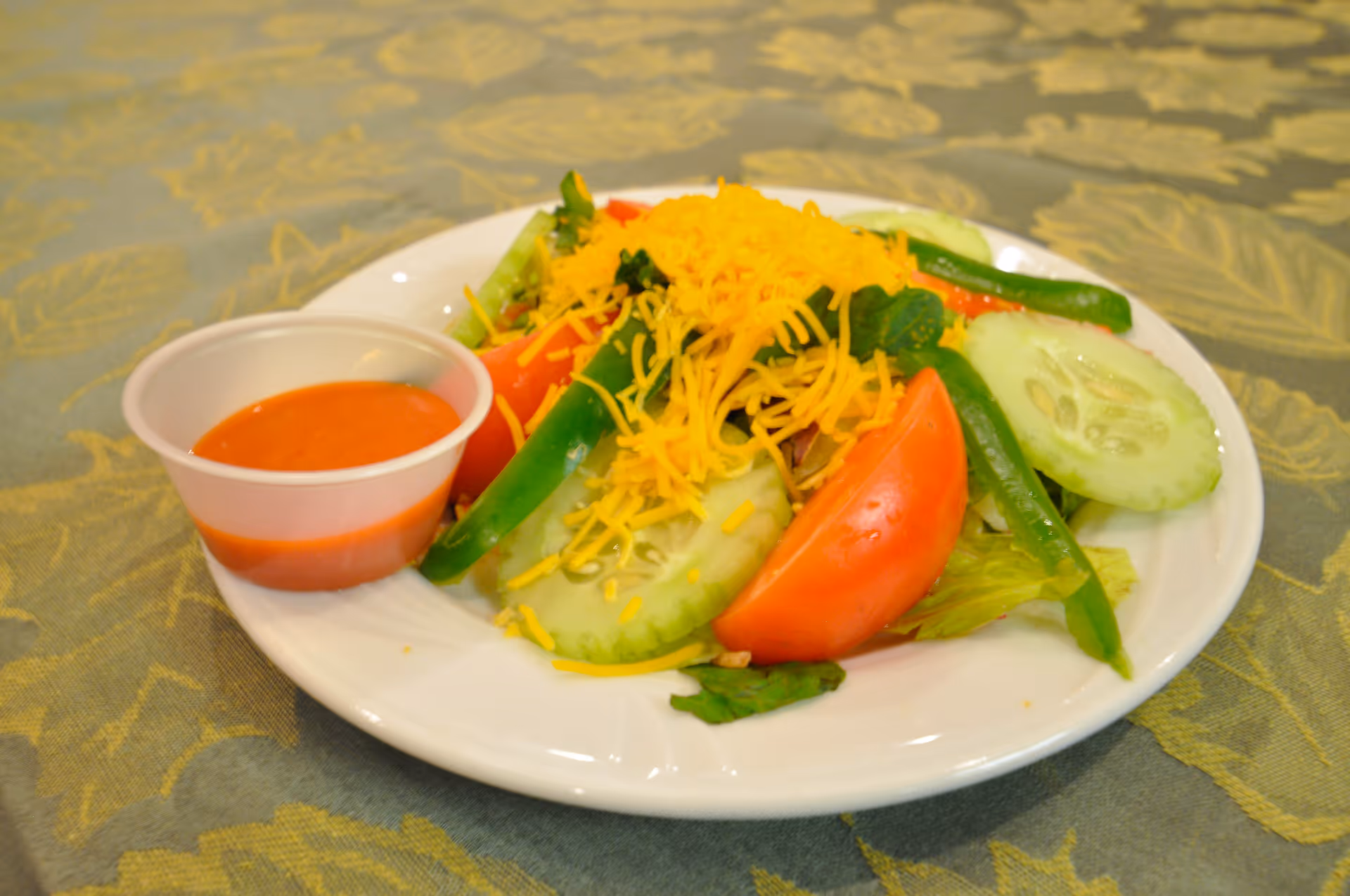 A white plate with a fresh salad consisting of sliced cucumbers, tomato wedges, green beans, lettuce, and shredded cheddar cheese, accompanied by a small cup of red dressing on the side, placed on a patterned tablecloth.