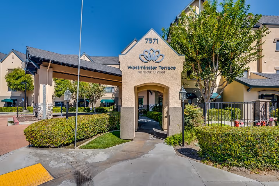 Entrance to Westminster Terrace Senior Living facility with a beige archway structure displaying the name and address number 7571. The building is surrounded by green bushes, trees, and a clear blue sky.