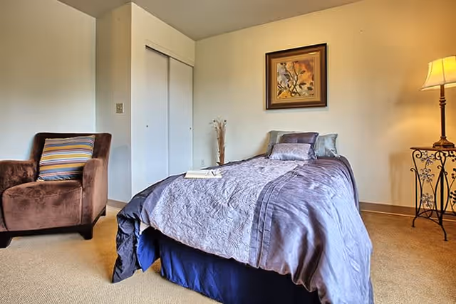 A cozy bedroom featuring a single bed with a purple quilt and multiple pillows, a brown armchair with a striped cushion, a framed floral painting on the wall, a standing lamp on an ornate metal side table, and a closed white closet door.
