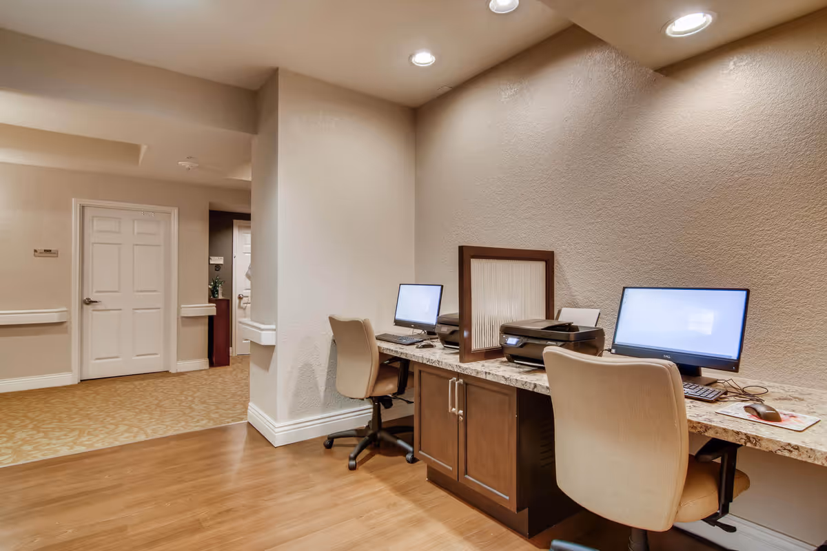 A small computer workstation area in a senior living facility with two desks, each equipped with a computer monitor, keyboard, mouse, and a printer. The desks have a marble-like countertop and dark wooden cabinets underneath. Two beige office chairs are positioned at the desks. The walls are light beige with recessed lighting overhead, and the floor is a combination of wood and patterned carpet. A hallway with closed white doors is visible in the background.