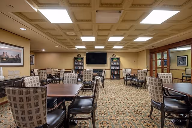 A spacious room with multiple square wooden tables surrounded by patterned cushioned chairs. The room has a patterned carpet floor, a coffered ceiling with recessed lighting, and beige walls. At the far end, there is a large flat-screen TV mounted on the wall flanked by two bookshelves filled with books. There are framed pictures on the walls and a sideboard with decorative items on the left side.