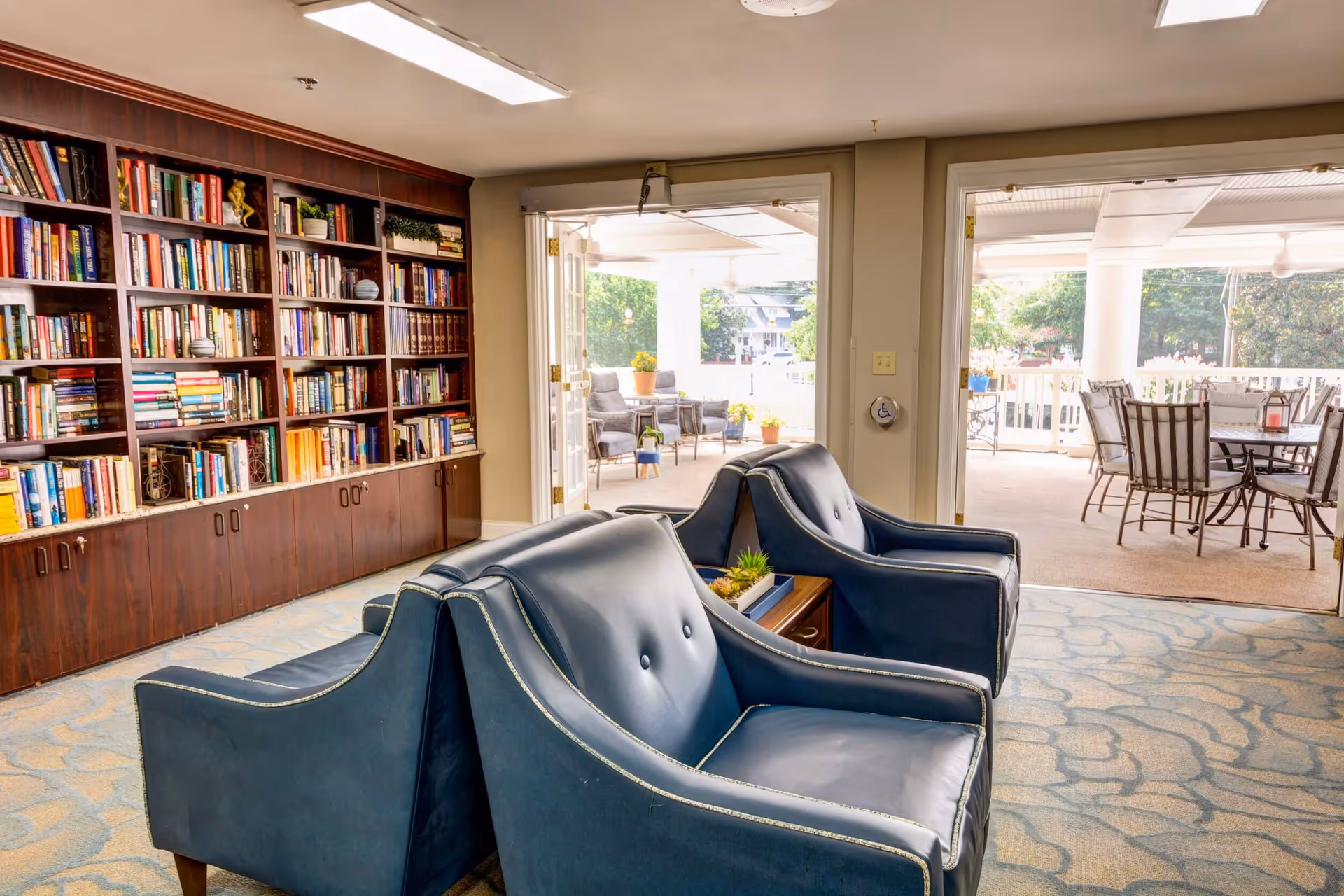Interior view of a cozy common area with two blue leather armchairs facing a large wooden bookshelf filled with books. The room opens up to an outdoor patio with seating and tables, surrounded by greenery.