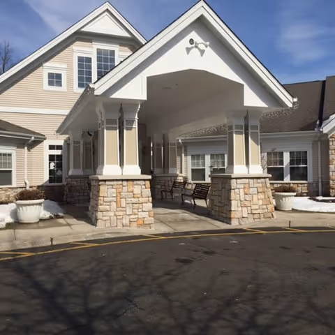 Entrance of a senior living facility with a covered drop-off area supported by stone and white columns. The building has beige siding, multiple windows, and a peaked roof. There are benches under the covered area and some snow on the ground nearby.