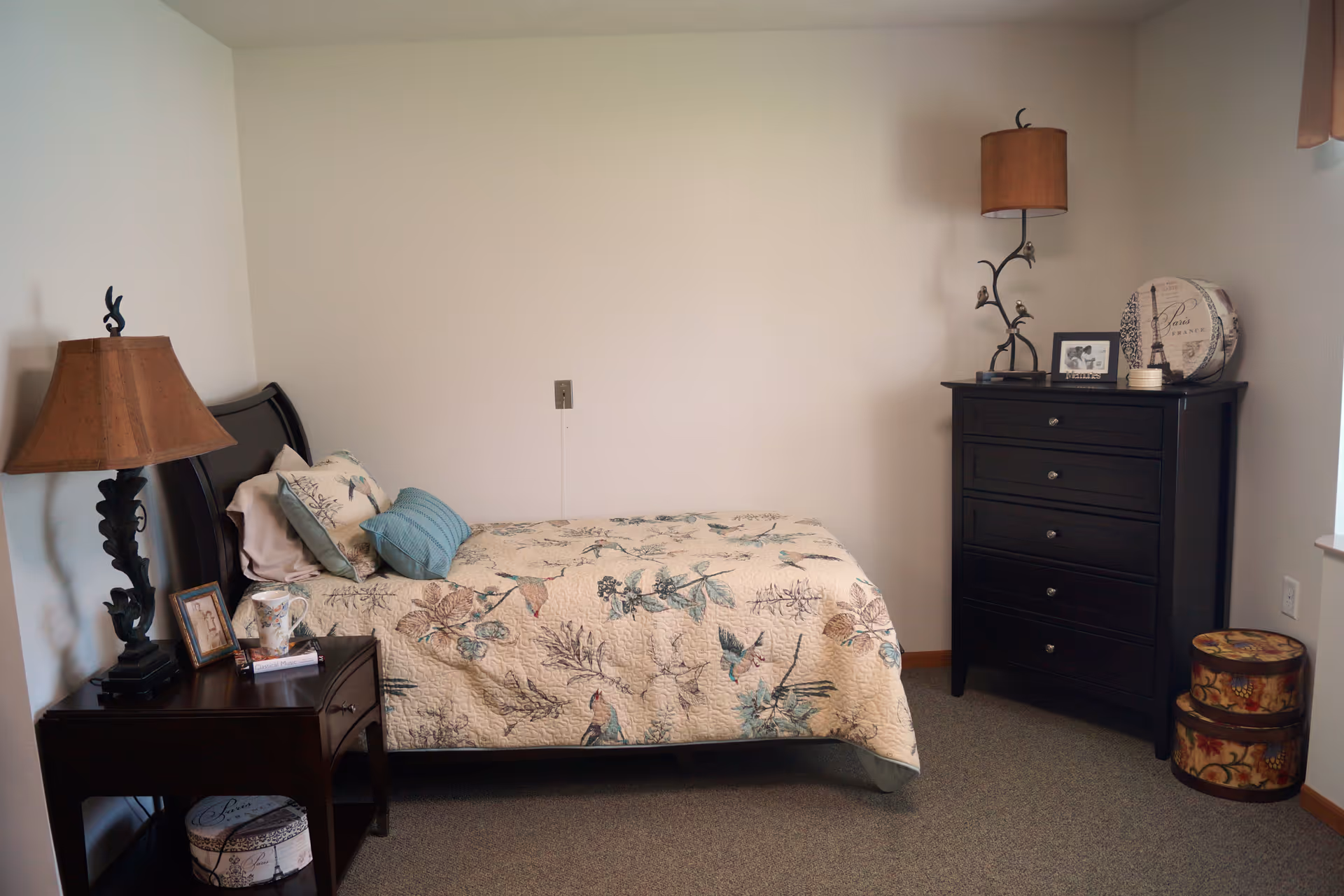 A small bedroom with a single bed covered in a floral patterned quilt. There is a dark wooden nightstand with a lamp, a framed photo, and a mug on it. Next to the bed is a tall dark wooden dresser with a decorative lamp, a framed photo, and round storage boxes on the floor beside it. The walls are plain and light-colored, and the floor is carpeted.
