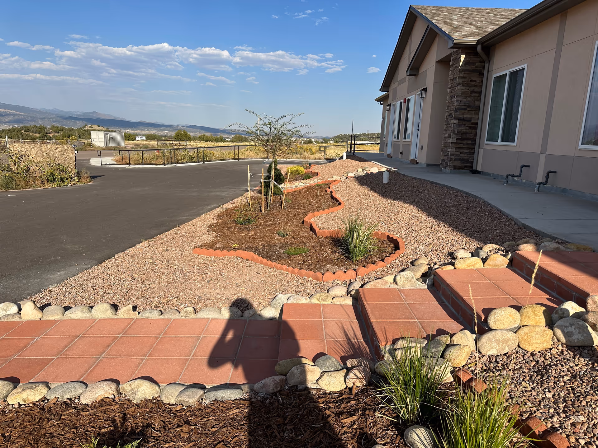 Outdoor view of a facility showing a paved walkway and steps bordered by rocks and landscaping with small plants and mulch. The building has beige walls with stone accents and several windows. In the background, there is a paved road, fencing, and a scenic view of distant hills under a partly cloudy blue sky.
