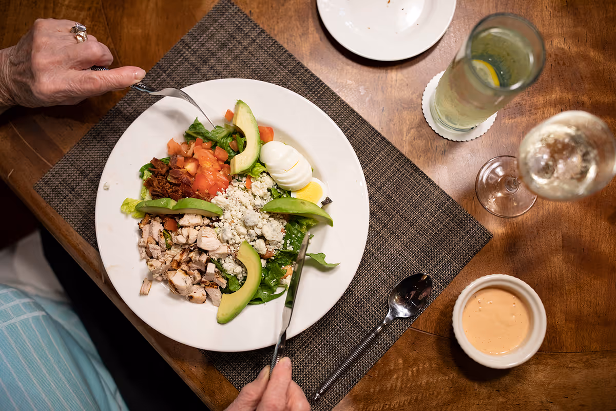 A person eating a salad with avocado slices, chopped chicken, tomatoes, bacon bits, blue cheese crumbles, and a hard-boiled egg on a white plate. The plate is on a brown woven placemat on a wooden table. There is a glass of water with lemon, a glass of white wine, a small bowl of dressing, and a spoon on the table.