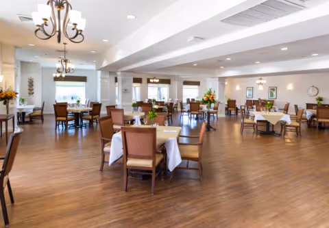 A spacious dining room with multiple tables covered with white tablecloths and set with chairs. The room has wooden flooring, large windows with blinds, and several chandeliers hanging from the ceiling. There are flower arrangements on some tables and framed artwork on the walls.