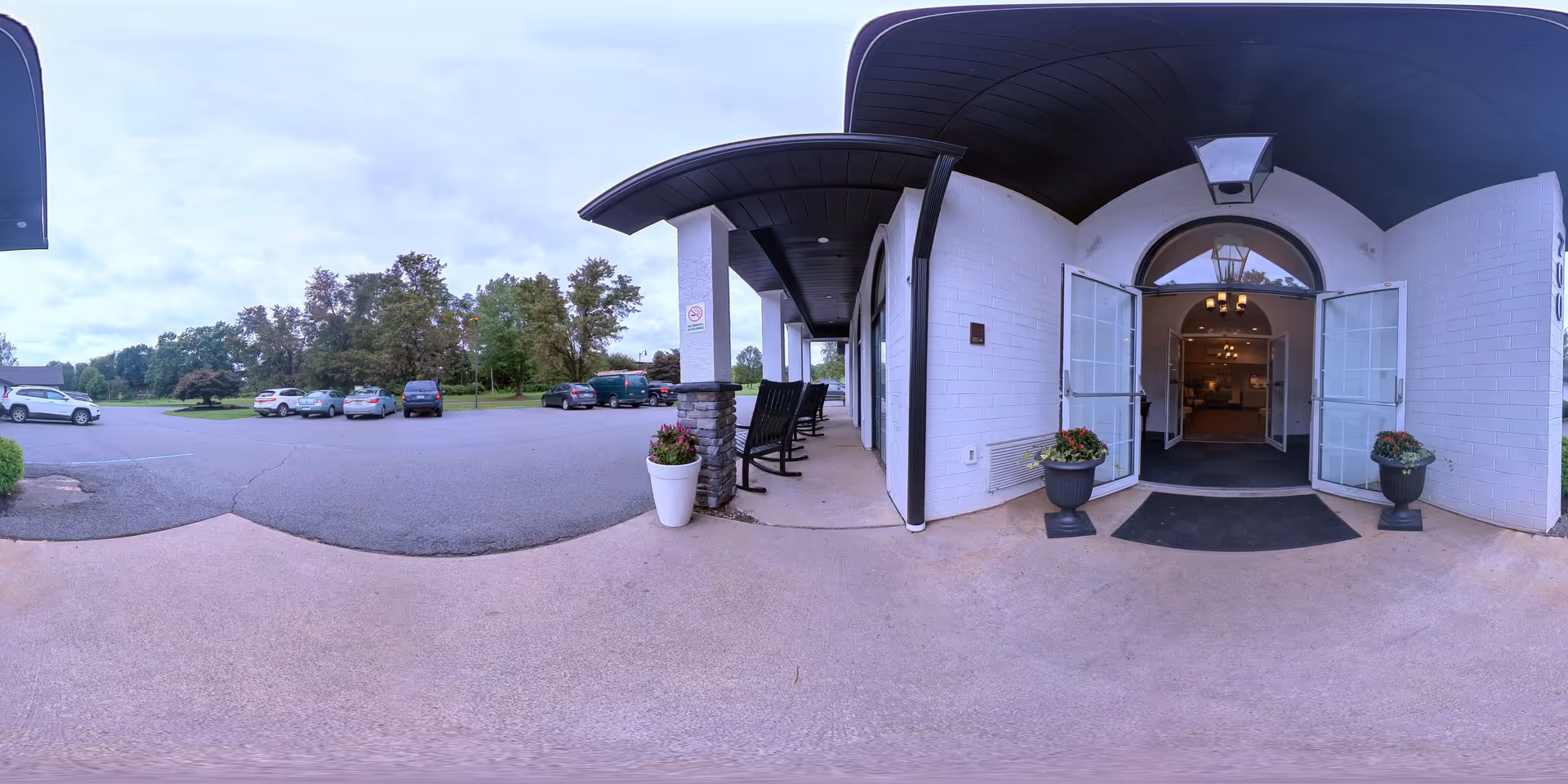 Wide-angle view of the entrance to Serenity Care Kingston facility showing an open double door entrance with two large planters on either side, a covered porch area with rocking chairs, a parking lot with several cars, and trees in the background under a cloudy sky.