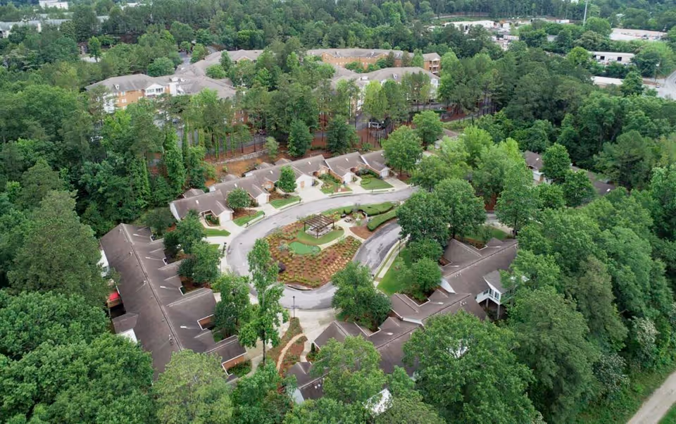 Aerial view of Galleria Woods senior living facility surrounded by dense green trees. The facility consists of multiple buildings arranged in a circular pattern with a landscaped garden and walking paths in the center. Additional buildings and parking areas are visible in the background.
