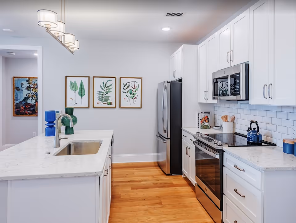 Bright modern kitchen with a marble island and sink, stainless steel refrigerator and stove, white cabinets, and three framed botanical prints on the wall.