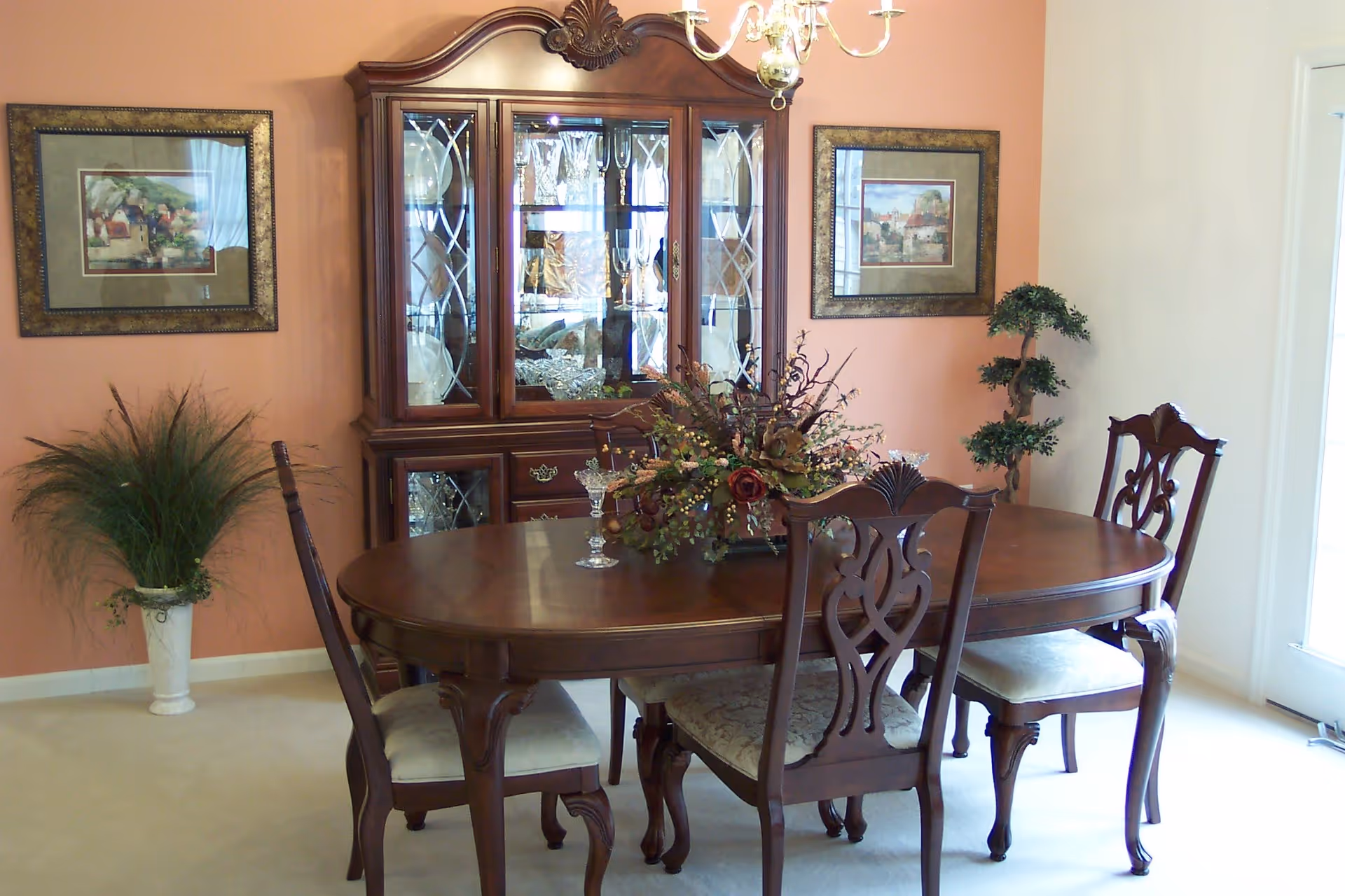A traditional dining room with a wooden oval dining table and four matching chairs with cushioned seats. A floral centerpiece and glassware are on the table. Behind the table is a wooden china cabinet with glass doors displaying crystal and glassware. The walls are painted in a warm peach color with two framed paintings and a potted plant in a white vase on the left side. A chandelier hangs above the table.