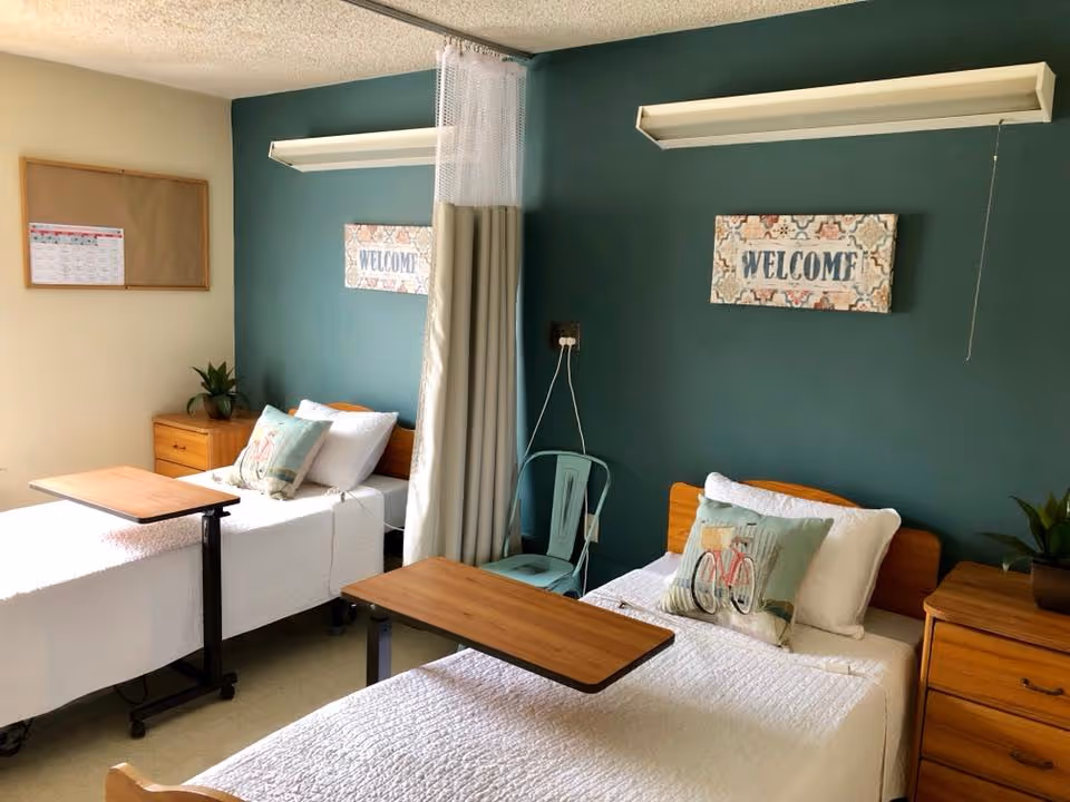A shared bedroom in a senior living facility with two single beds separated by a privacy curtain. Each bed has white bedding, a decorative pillow, and an overbed table. The walls are painted teal, and there are two wooden nightstands with small plants. Above each bed is a decorative sign that says 'WELCOME'. A metal chair is placed between the beds.