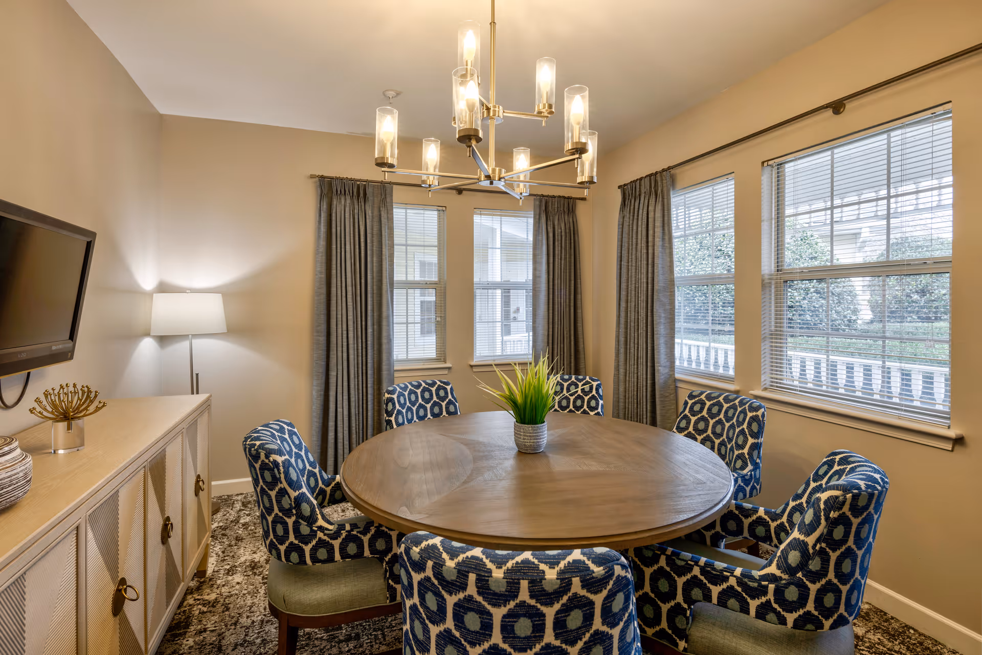 A dining room with a round wooden table surrounded by six blue patterned chairs. There is a modern chandelier hanging above the table and a small potted plant in the center. The room has three windows with gray curtains and blinds, a floor lamp, a wall-mounted TV, and a sideboard with decorative items.