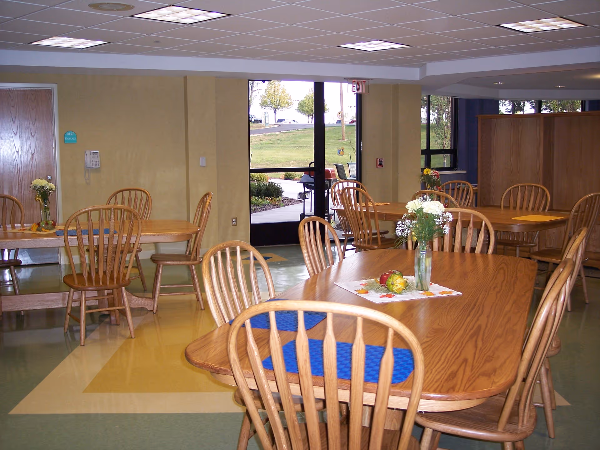 Interior view of a dining room with several wooden tables and chairs. Each table has a small floral centerpiece and placemats. There is a glass door leading outside, and the room has a tiled floor with a geometric pattern. The walls are light-colored, and there is a wooden cabinet in the background.