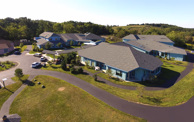 Aerial view of a senior living facility named Camphill Ghent, showing multiple single-story buildings with gray roofs surrounded by green lawns, trees, and paved pathways. Several cars are parked near the buildings, and the area is set against a backdrop of wooded hills under a clear blue sky.