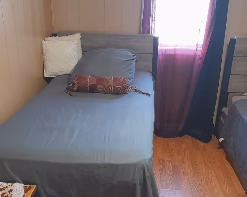 A bedroom with two single beds featuring gray headboards and gray bedding. One bed has a white decorative pillow and a patterned cylindrical pillow. There is a window with sheer purple and black curtains letting in light, and the floor is wooden.