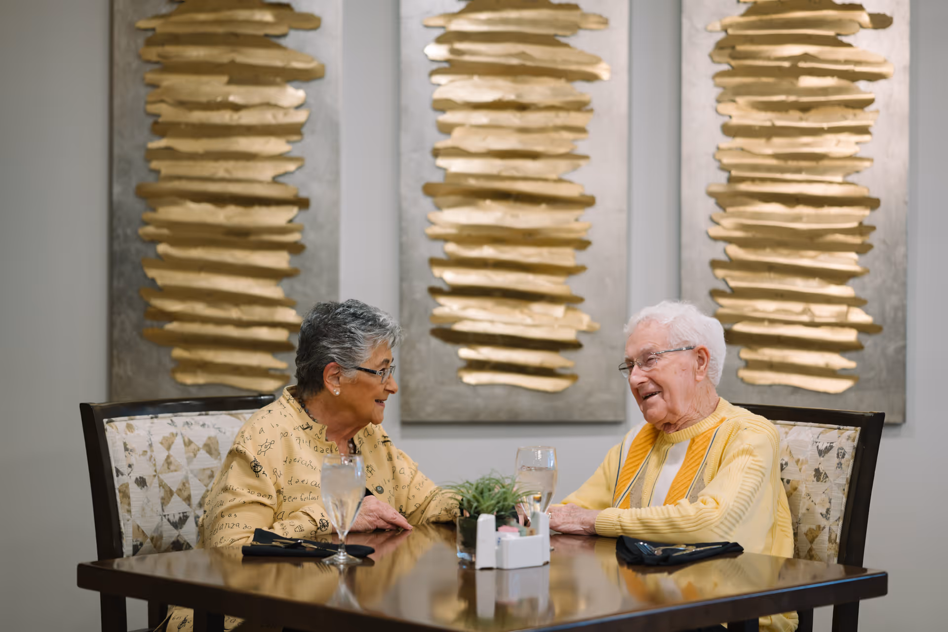 Two elderly individuals sitting at a dining table in a senior living facility, engaged in conversation. The table has two glasses of water, black napkins, and a small plant centerpiece. Behind them are three large decorative wall art pieces with gold and silver abstract designs.