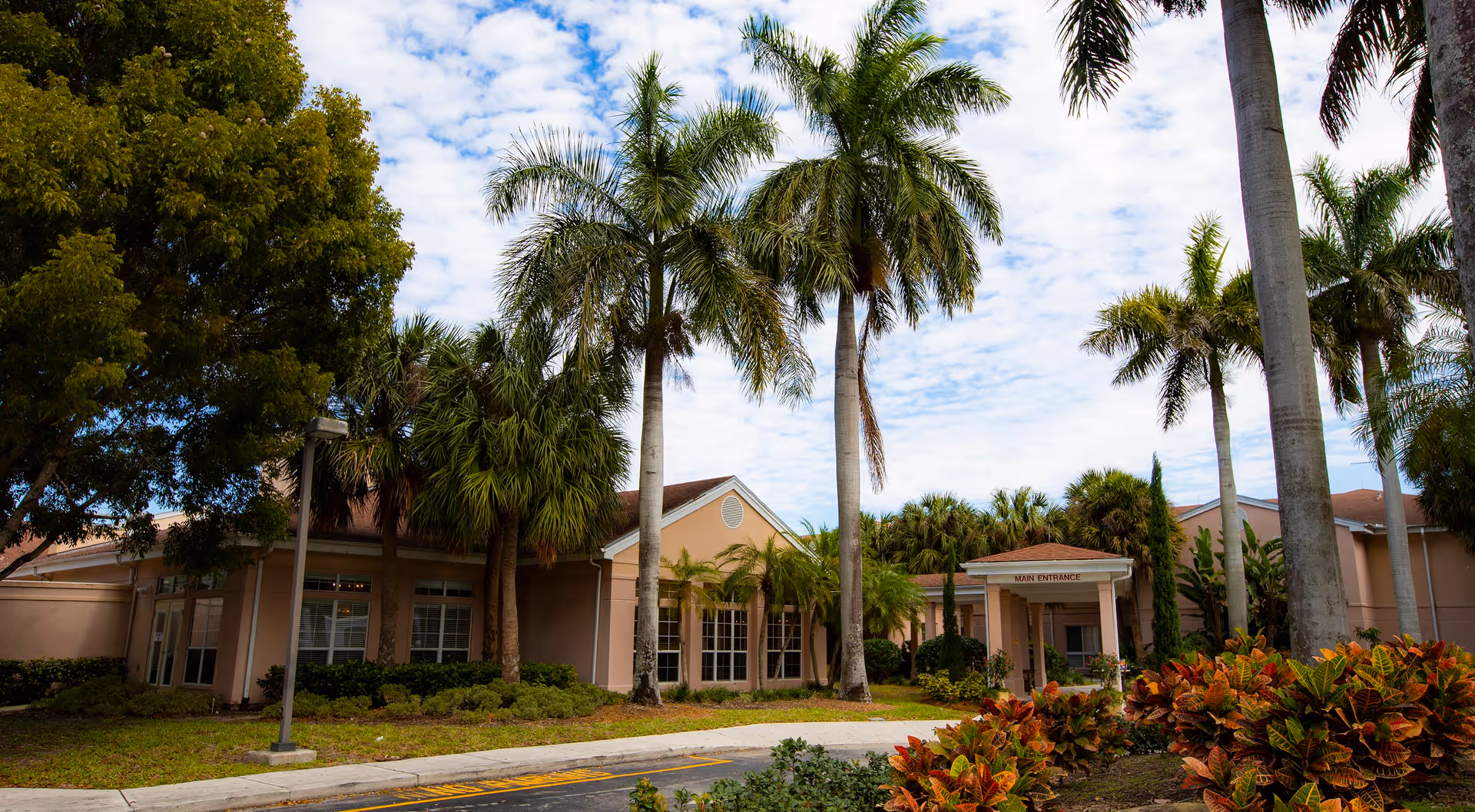 Exterior view of a single-story pink building entrance surrounded by palm trees and landscaped shrubs under a partly cloudy sky.