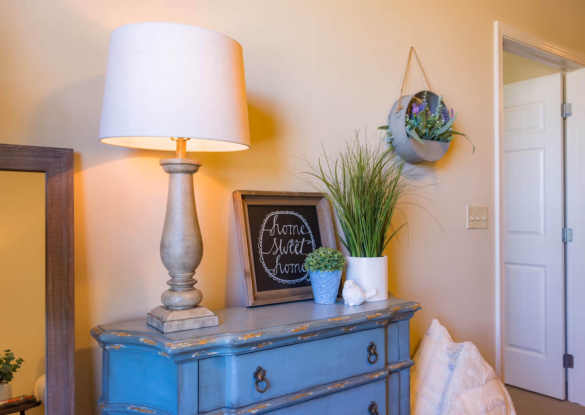 A cozy corner of a room featuring a distressed blue wooden dresser with a beige table lamp, a framed chalkboard sign that reads 'home sweet home', two potted plants, and a small white bird figurine. A round wall planter with flowers hangs on the beige wall above the dresser, and a white door is partially open in the background.