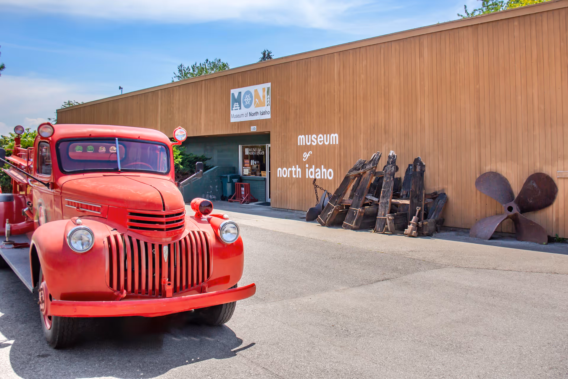 A vintage red fire truck parked outside a wooden building with a sign that reads 'museum of north idaho' and 'Museum of North Idaho'. Various large wooden and metal artifacts are displayed against the building's exterior wall.
