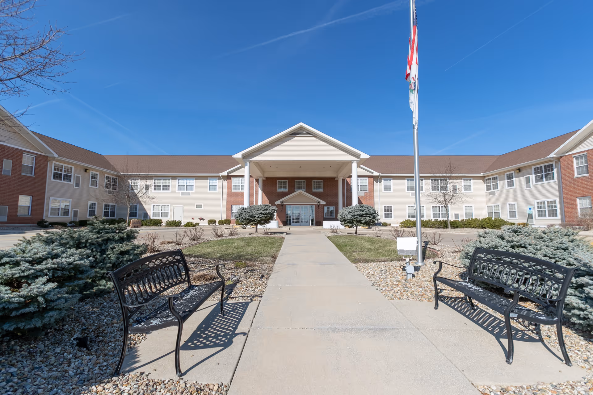 Front entrance of a two-story senior living building with a covered portico, walkway, flagpole, benches, and landscaping.