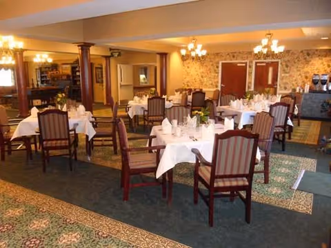 A dining room with multiple tables covered in white tablecloths, each set with plates, cups, and napkins. The room has patterned carpet, wooden chairs with striped upholstery, chandeliers hanging from the ceiling, and floral wallpaper on the far wall.