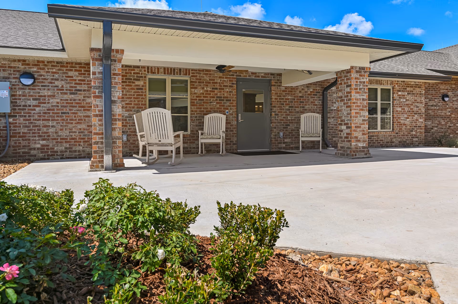 Exterior view of a brick building with a covered porch area featuring three beige rocking chairs. There are two windows and a gray door under the porch roof. In the foreground, there are green bushes and flowers planted in a mulched garden bed. The sky is blue with some clouds.