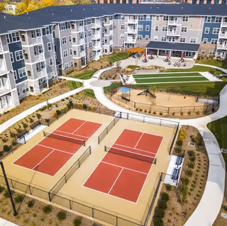 Aerial view of Encore 55+ Boutique Apartments - Northeast showing a multi-story residential building surrounding an outdoor recreational area with two red pickleball courts, a playground, walking paths, and landscaped greenery.