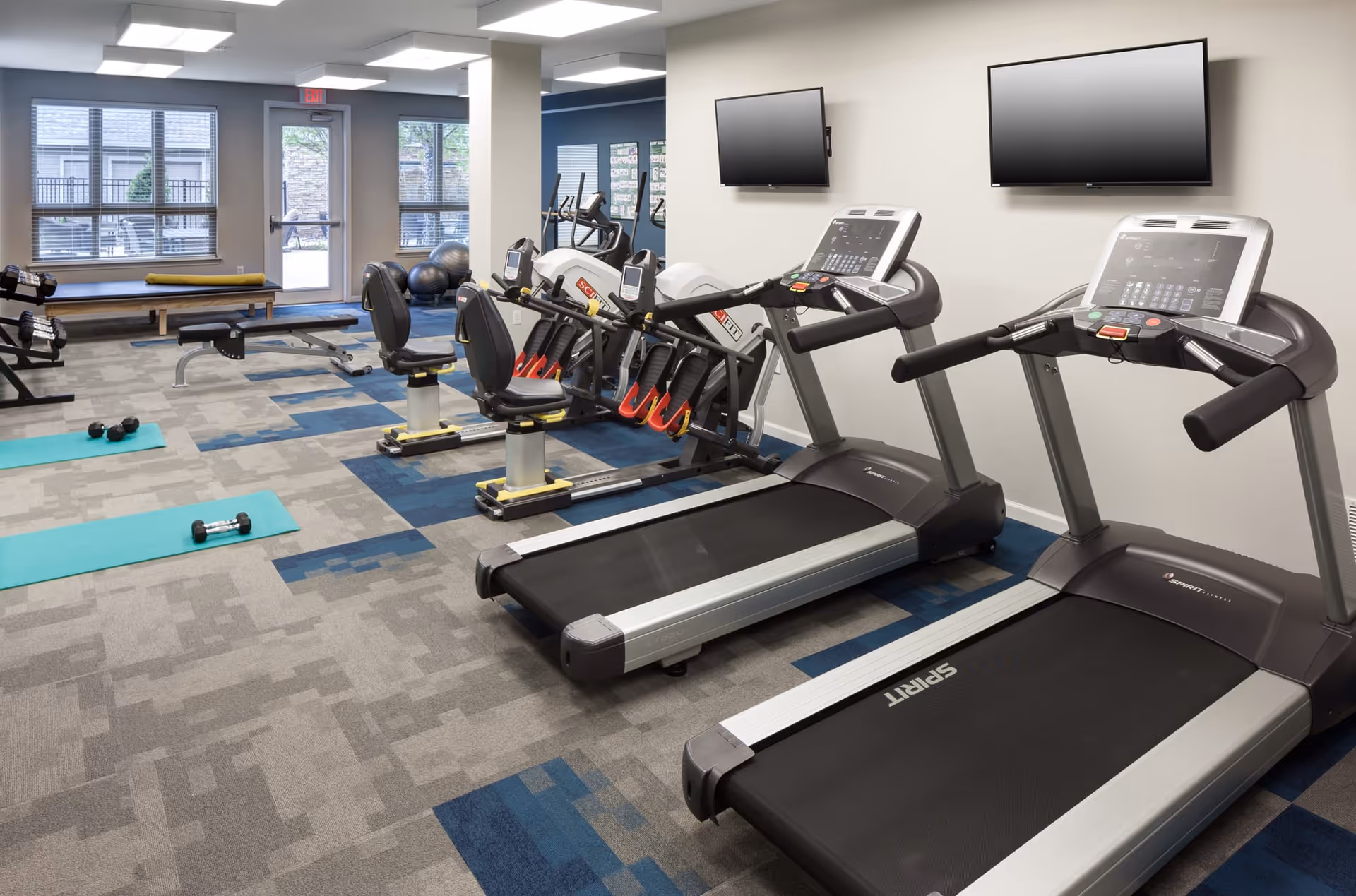 A fitness room with two treadmills, two recumbent exercise bikes, a rowing machine, dumbbells, exercise mats, and large windows letting in natural light.