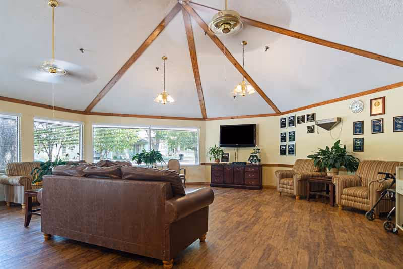 A spacious living room area with a high ceiling featuring wooden beams and ceiling fans. The room has large windows letting in natural light and showing greenery outside. Inside, there is a brown leather sofa, several striped armchairs, a wooden cabinet with a flat-screen TV mounted above it, and various plants and plaques on the walls. The floor is wooden, and the overall atmosphere is warm and inviting.