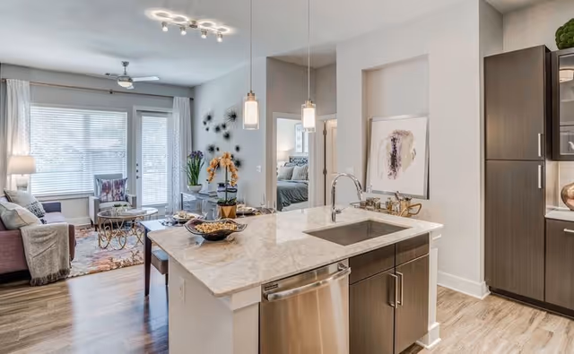 Modern open-concept living space featuring a kitchen island with a marble countertop and built-in sink, pendant lights hanging above, dark wood cabinets, and stainless steel dishwasher. In the background, there is a living room area with a sofa, armchair, coffee table, and large windows with blinds allowing natural light to fill the room. A bedroom is partially visible through an open doorway.