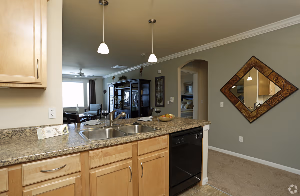 View of a kitchen area with a double sink and granite countertop, light wood cabinets, and a black dishwasher. Two pendant lights hang from the ceiling above the counter. In the background, there is a living room with a large window, a black entertainment center, chairs, and a ceiling fan. To the right, a hallway leads to a bedroom with a bed visible. A decorative diamond-shaped mirror hangs on the wall near the hallway.