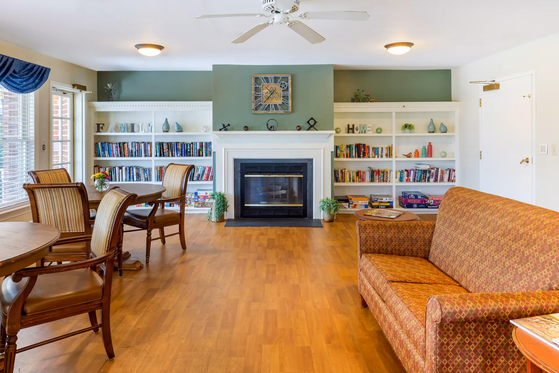 A cozy common area in a senior living facility featuring a fireplace centered between two white bookshelves filled with books, board games, and decorative items. There are several wooden chairs with striped cushions around round tables on the left side, and a patterned upholstered sofa on the right. The room has wood flooring, a ceiling fan, and soft lighting.