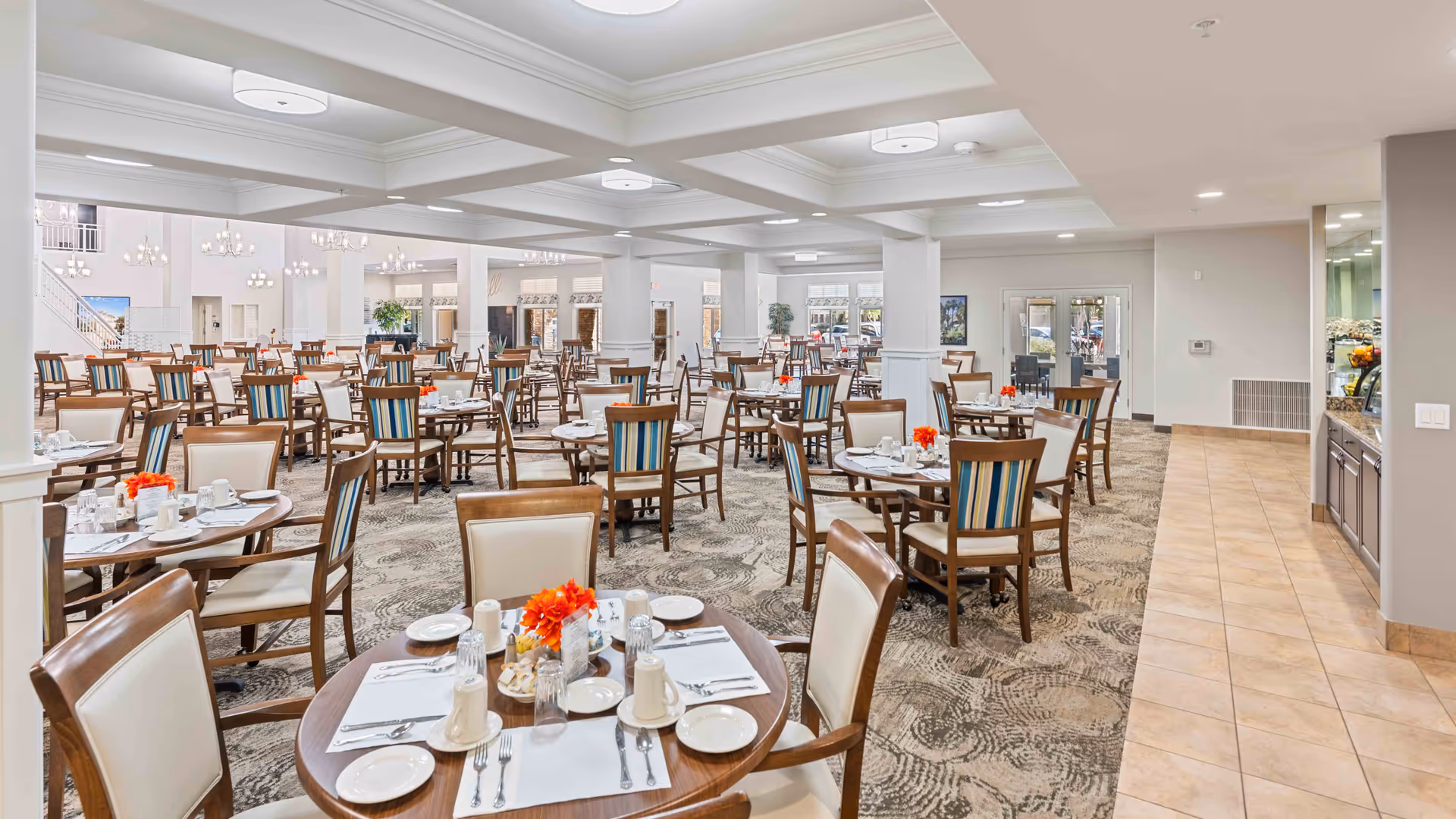Spacious dining room in a retirement living facility with multiple round tables set with white plates, cups, silverware, and small orange flower centerpieces. The room features carpeted flooring with a patterned design, beige walls, and ceiling lights. There are wooden chairs with cream-colored cushions and some with blue-striped backs. Large windows and glass doors allow natural light to enter the room.