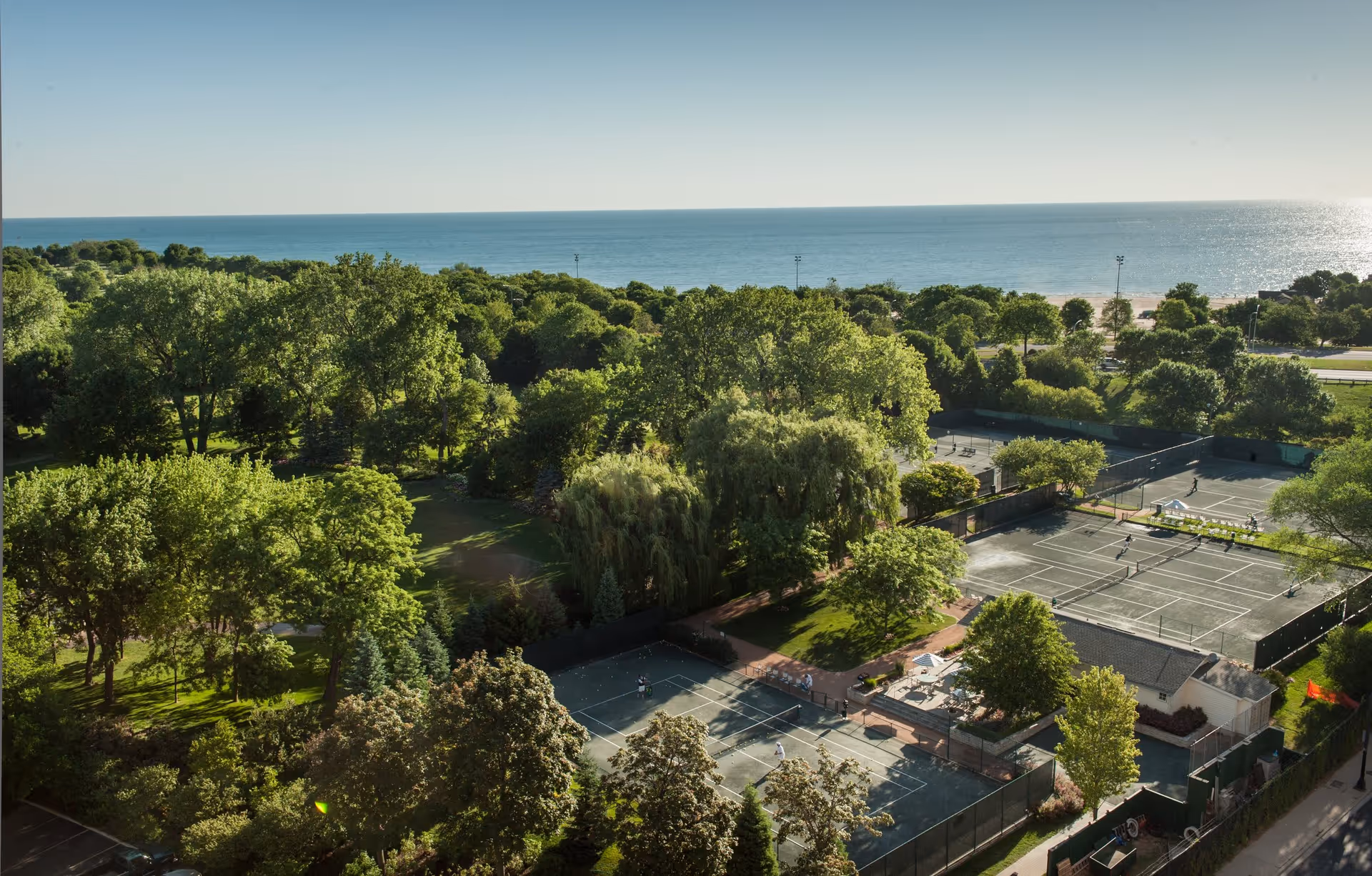 Aerial view of a green park area with many trees, tennis courts, and a large body of water in the background under a clear sky.