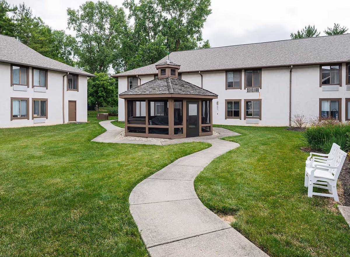 A well-maintained outdoor courtyard area at The Village of Westerville featuring a curved concrete walkway leading to a screened gazebo. The courtyard is surrounded by two-story residential buildings with multiple windows. There are green lawns, trees in the background, and white benches along the walkway.