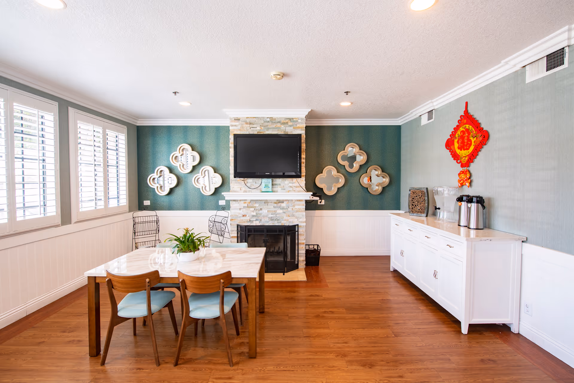 Bright dining area with a wooden table and chairs, a fireplace with a mounted TV, decorative mirrors on a teal wall, and a white sideboard with coffee urns.