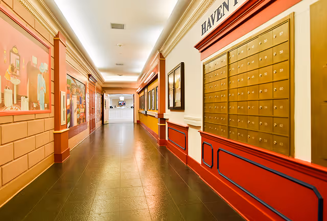 A long, brightly lit decorated hallway with wall-mounted mailboxes, framed artwork, and a reception desk at the far end.