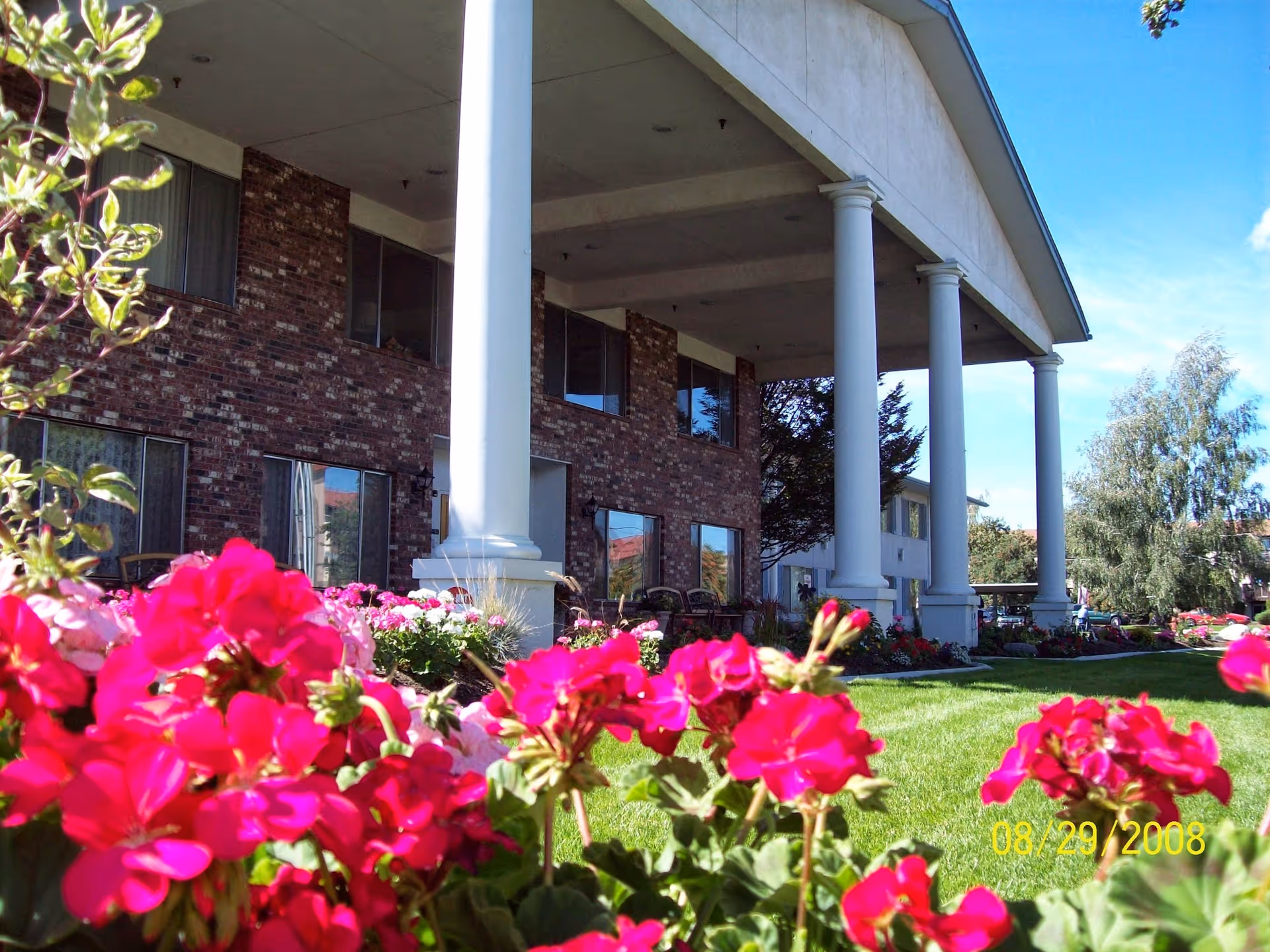 Front entrance of a brick senior living building with tall white columns and pink flowers in the foreground.