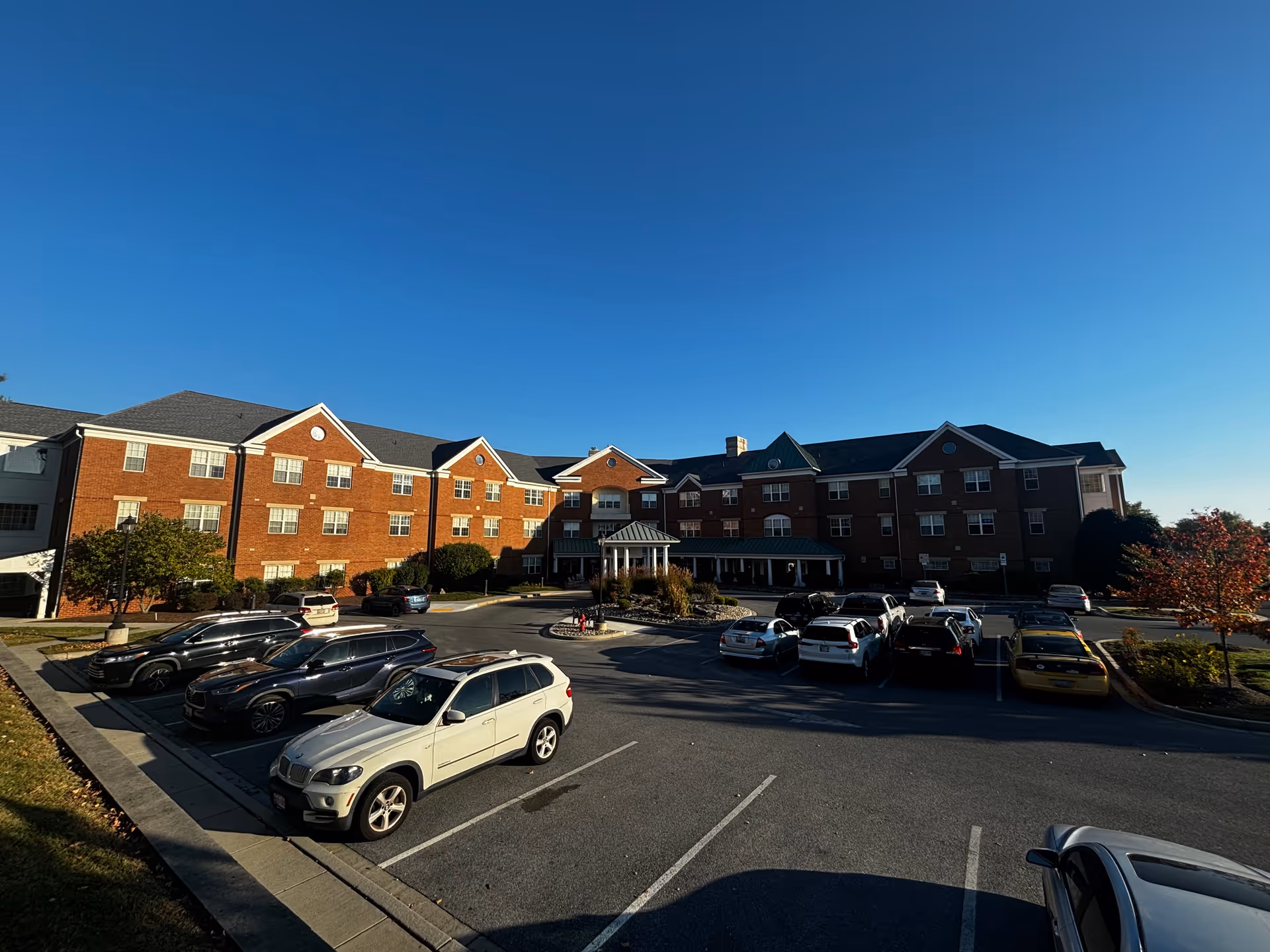 Exterior view of a large three-story brick building under a clear blue sky with a parking lot in front containing several parked cars. The building has multiple windows and a covered entrance with a small gazebo-like structure in the center.