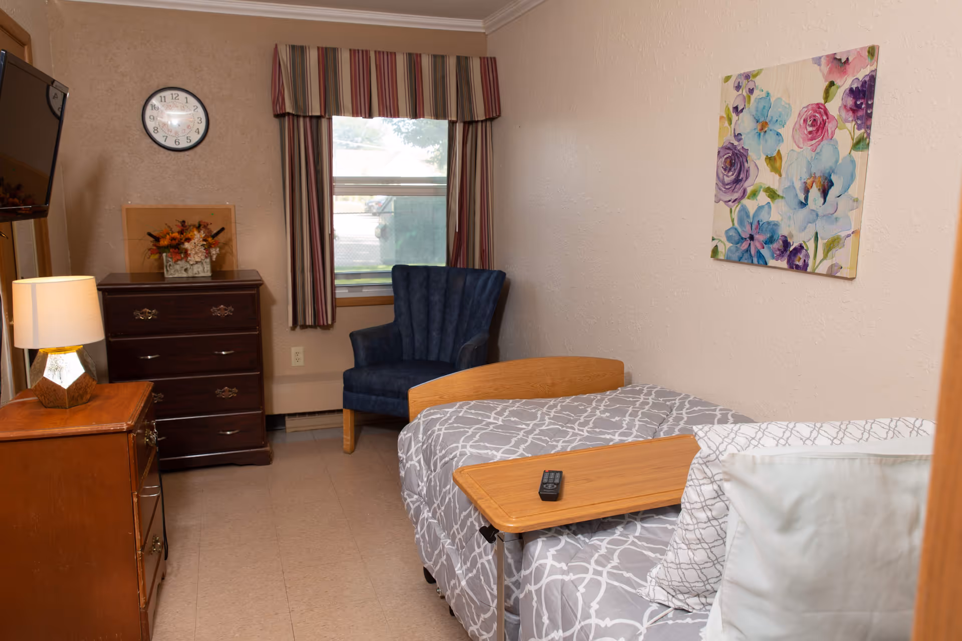 A small bedroom with a single bed covered in gray and white patterned bedding. There is a wooden overbed table with a remote control on it. A blue upholstered armchair is placed near a window with striped curtains. A dark wooden dresser with a floral arrangement and a wall clock above it is on the left side. A colorful floral painting hangs on the right wall. A lamp sits on a wooden nightstand beside the bed.