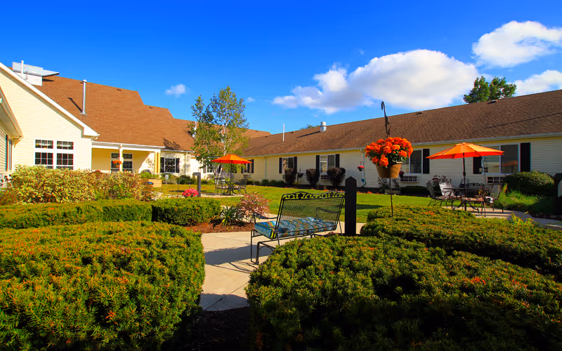 Landscaped outdoor courtyard with benches, patio tables and orange umbrellas in front of single-story facility buildings.