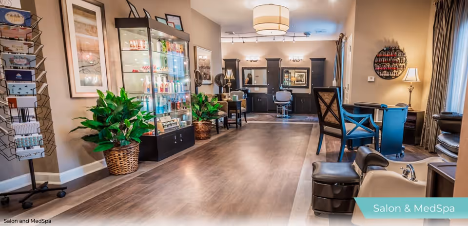 Interior view of a salon and medspa area with wooden flooring, a display cabinet filled with beauty products, potted plants, salon chairs, mirrors, and manicure stations. The space is well-lit with ceiling lights and has a cozy, welcoming atmosphere.