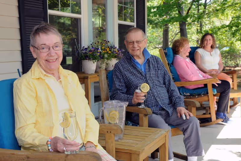 Four people sitting on wooden rocking chairs on a porch. Two elderly individuals in the foreground are holding glasses of lemonade, with a pitcher of lemonade on a small table between them. Two women are seated further back, smiling and enjoying the outdoor setting surrounded by trees and greenery.