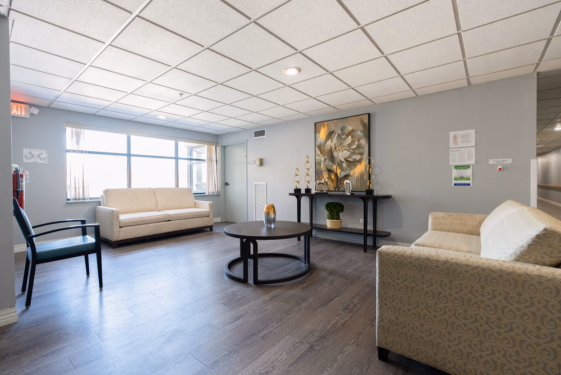 A bright and clean supportive living facility common area with two beige sofas, a black armchair, a round black coffee table with a decorative vase, a console table with decorative items and a large floral painting on the wall. The room has large windows letting in natural light, light gray walls, and wood-look flooring.