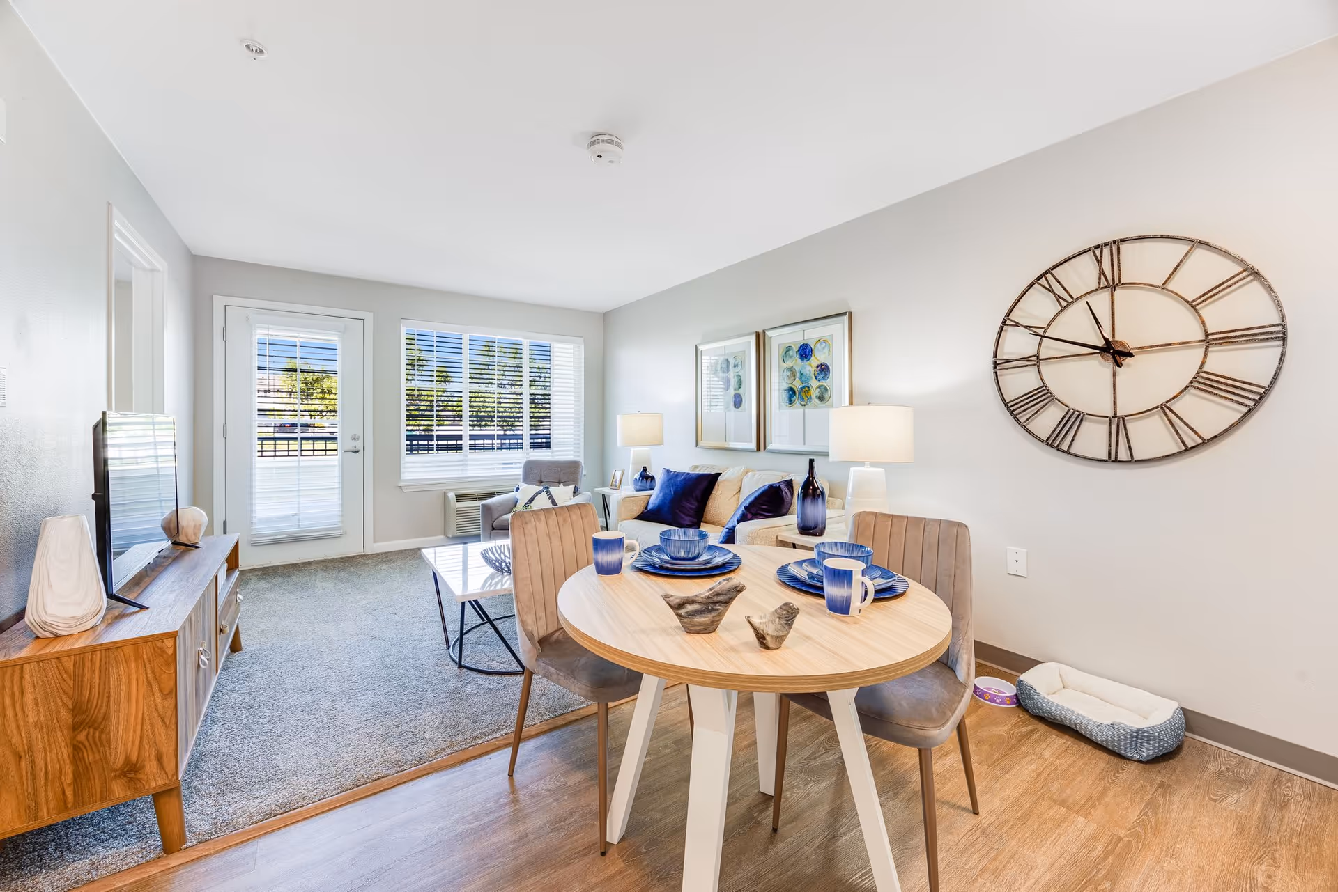 A bright and modern living room and dining area with a round wooden dining table set for two with blue dishes and mugs. The living room has a beige couch with blue pillows, a coffee table, a gray armchair, two table lamps, and two framed artworks on the wall. A large decorative clock hangs on the wall near a pet bed and food bowl. There is a wooden TV stand with a flat-screen TV and decorative items. Large windows and a glass door with white blinds allow natural light to fill the room.