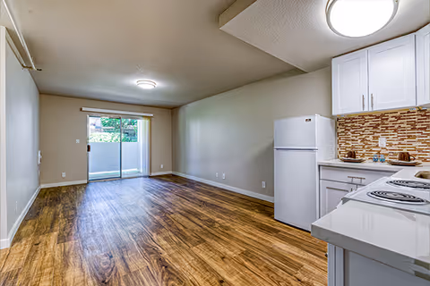 Interior view of a senior apartment unit showing a small kitchen area with white cabinets, a refrigerator, and an electric stove on the right. The room has wood-style flooring and beige walls. At the far end, there is a sliding glass door leading to an outdoor patio or balcony with greenery visible outside.