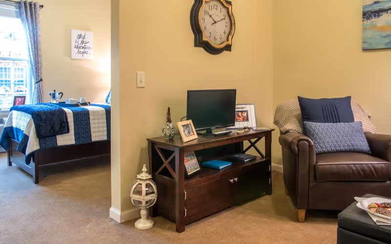 A cozy living space featuring a brown leather armchair with two pillows, a wooden TV stand with a small flat-screen television, framed photos, and decorative items. To the left, a bedroom is partially visible with a bed covered in a blue and white quilt, a window with curtains, and a wall decoration with text. A decorative clock hangs on the wall above the TV stand.