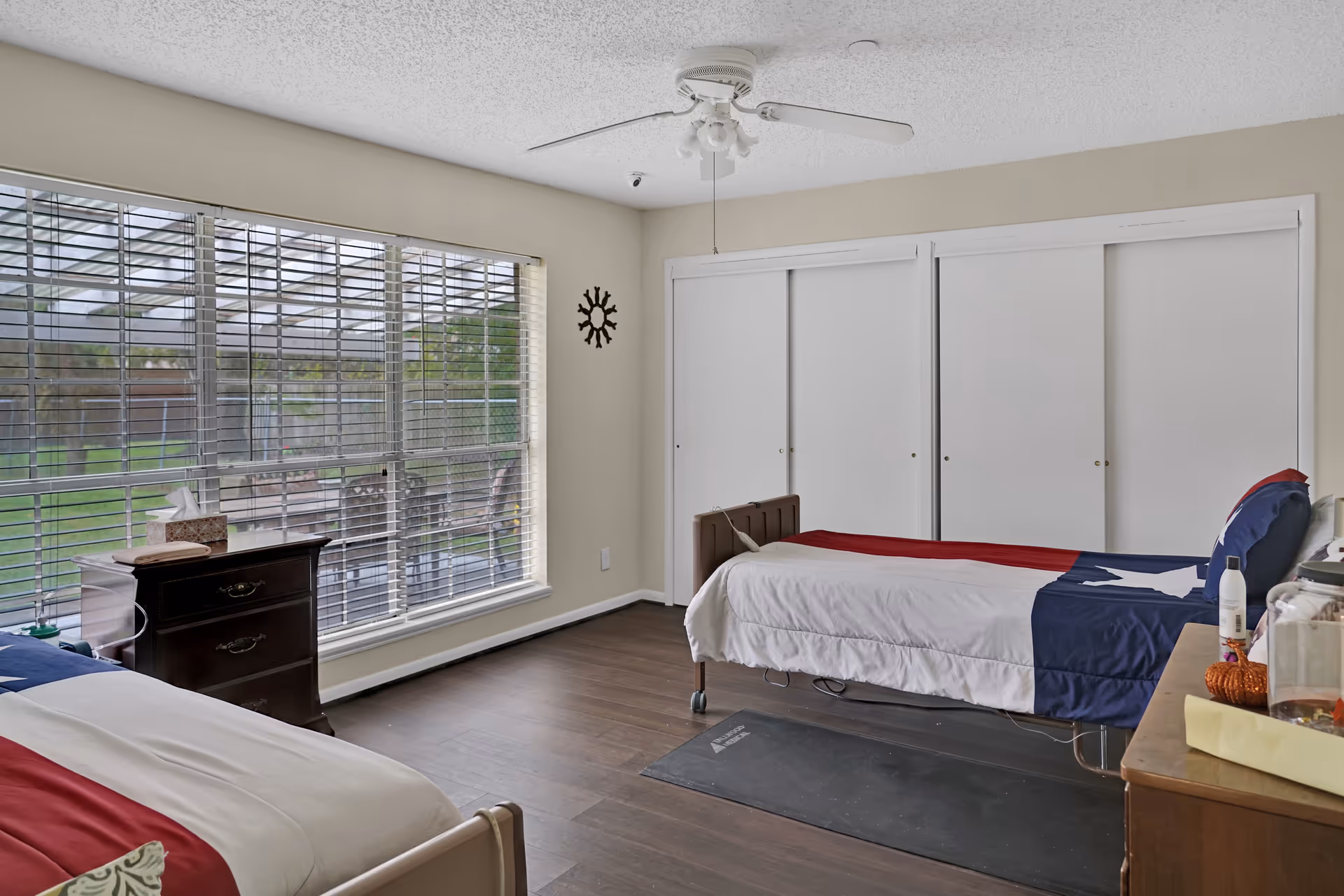 A bright bedroom with two single beds, each with a patriotic-themed bedspread. The room has large windows with blinds allowing natural light to enter, a ceiling fan, wooden flooring, and a set of white sliding closet doors. There are bedside tables with various items on them.